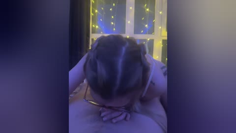 Video of a person with braided hair, topless, standing in front of a window with glowing yellow fairy lights, hands on their head, viewed from behind.