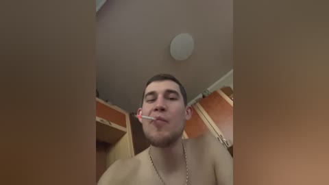 Video of a young man with short dark hair, no shirt, holding a lit cigarette to his mouth, standing in a beige room with wooden cabinets and a ceiling light.