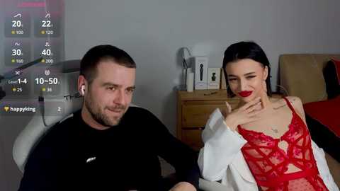 Video of a man and woman in a bedroom. Man has short brown hair, beard, black shirt; woman with dark hair, red lace lingerie, white robe, smiling. Room has wooden dresser, white walls, digital clock.