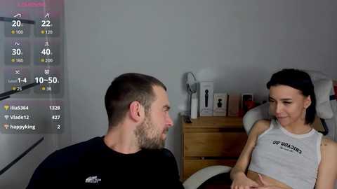 Video of a bearded man in black shirt and a woman in a white tank top, both smiling, in a modern bedroom with a wooden nightstand and a white chair.
