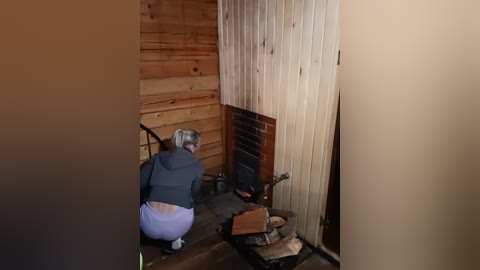 A video of a person with short gray hair in a gray hoodie and white pants kneeling beside a small wood stove in a rustic room with wooden walls and a brick hearth.