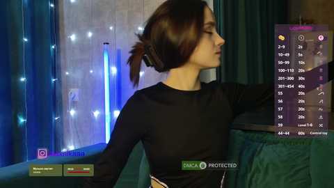 Video of a slender woman with fair skin and dark brown hair in a bun, wearing a black long-sleeve top, standing by a glowing blue LED wall, with a digital app interface in the foreground.