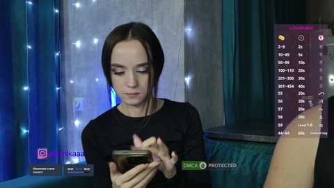 A video of a young woman with fair skin and straight brown hair, wearing a black top, intensely focused on her smartphone, surrounded by blue fairy lights in a dimly lit room.