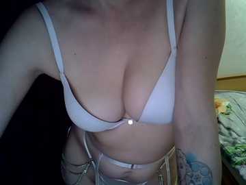 A close-up video of a woman's torso, wearing a white bra with a central bow and a beige belt with a chain detail. She has a tattoo on her left arm and is in a dimly lit bedroom.
