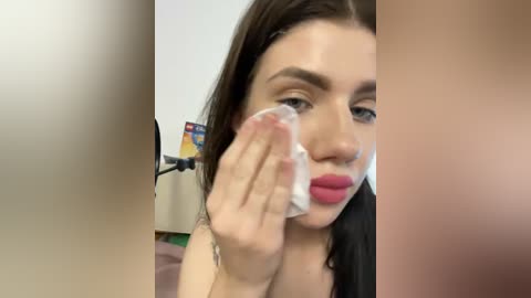 Video of a young woman with fair skin, dark hair, and bright red lipstick, wiping her face with a tissue. Background shows a blurred wall and a chair.