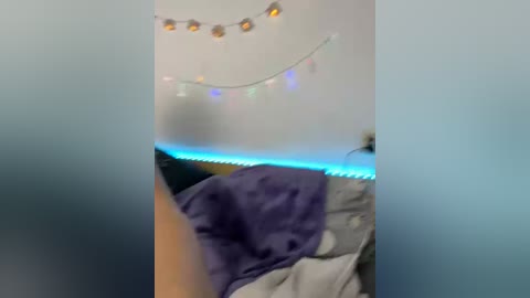 A blurry video of a bed with a white comforter and a purple blanket, adorned with a string of multicolored lights and a necklace, set against a white wall.