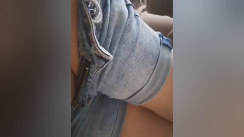 Video of a person's upper thigh and hip, wearing light blue denim shorts with a slight crease, sitting on a tan surface, blurred background.