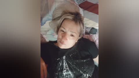 A video of a young woman with short, blonde hair, lying on a colorful bedspread, wearing a black shirt. The image is slightly blurred, with a soft focus.