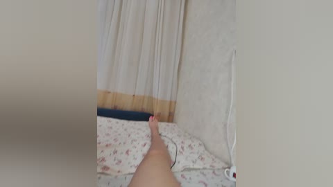 Video of a person lying on a bed with white and floral-patterned sheets, beige walls, and a white curtain partially drawn. The person's legs are stretched out, and a white electrical cord is visible.
