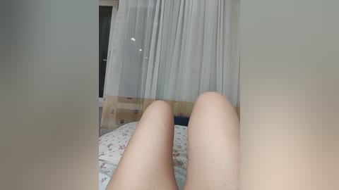 A video of a pair of light-skinned, slender legs lying on a bed with floral-patterned sheets, partially covered by sheer white curtains. The background reveals a softly lit room with wooden furniture.
