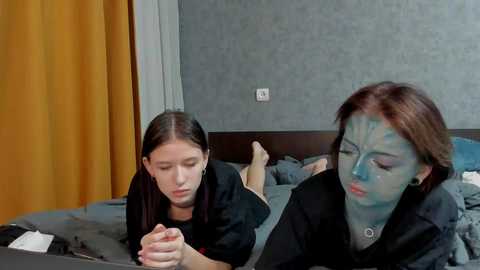 A video of a young woman with dark hair and a child with light skin and straight hair, both wearing black t-shirts, lying on a bed in a modern room with gray walls, a yellow curtain, and a wooden headboard.