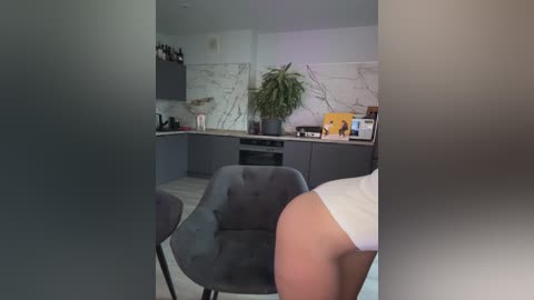 Video of a modern kitchen with grey cabinets, white marble backsplash, and a grey upholstered chair. A person in white underwear stands in the foreground, partially visible.