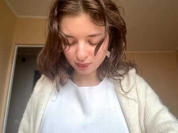 Video of a young woman with shoulder-length, wavy brown hair, wearing a white ribbed shirt under a beige cardigan, standing in a beige-walled room with a doorway in the background.