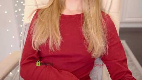 Video of a woman with long blonde hair wearing a red sweater, seated in a beige chair with a green key attached to the sweater.