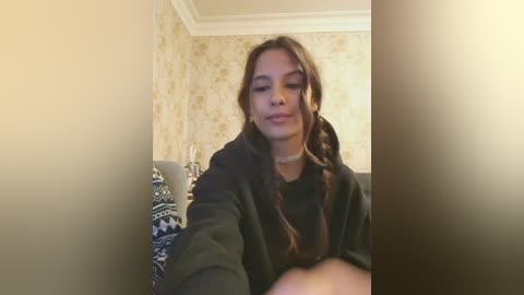 Video of a young woman with long brown hair, wearing a black coat, sitting on a patterned cushion in a cozy, dimly lit room with beige floral wallpaper.