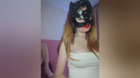 Video of a young, light-skinned woman with long, blonde hair, wearing a black leather cat mask, red lipstick, and a white, tight-fitting top. She's seated on a pink sofa in a dimly lit room.