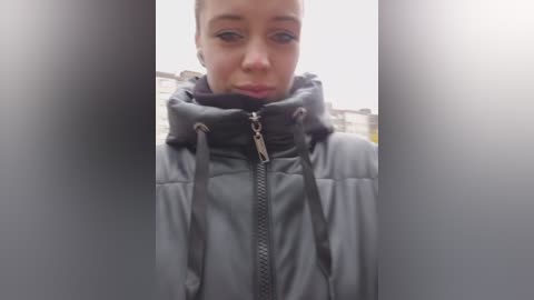 Video of a young woman with light skin, wearing a puffy, grey winter jacket, standing outdoors with blurred city buildings in the background.
