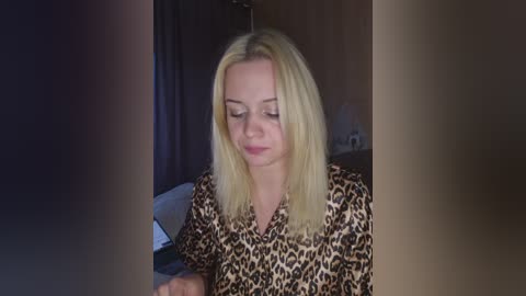 Video of a young Caucasian woman with shoulder-length blonde hair, wearing a leopard print blouse, seated indoors, focusing on a tablet, with dark curtains and dim lighting in the background.