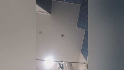 Video of a modern bathroom ceiling with two recessed lights, a silver shower rod, and blue-tiled corner accent. The walls are white, and the overall style is minimalist and clean.