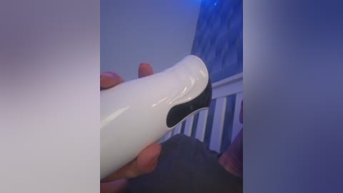 Video of a person's hand holding a white, sleek, black-tipped wand vibrator, with a blurred background showing a blue-lit staircase and a dark wooden floor.