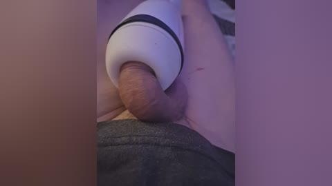 Video of a close-up view of a person's penis being stimulated by a white, cylindrical vibrator. The background is blurred, featuring a gray fabric surface. The image is intimate and explicit.