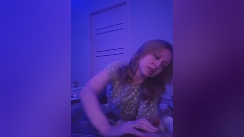 A video of a woman with long brown hair in a shimmering gold dress, leaning against a white door in a dimly lit room with purple lighting.