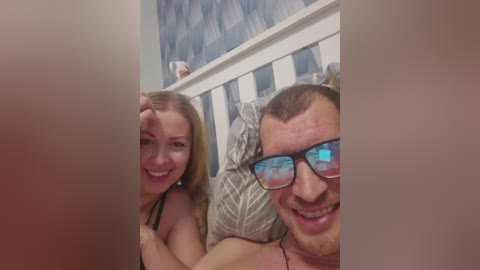 Video of a smiling couple in bed: a blonde woman with medium-sized breasts, and a man with short brown hair and dark glasses.