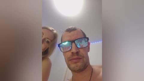A video shows a man with glasses and a woman with blonde hair smiling, both topless, in a brightly lit room with a round light fixture.