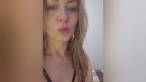 Video of a Caucasian woman with light skin and long, straight blonde hair. She is wearing a black bra with a patterned lace trim, and her lips are slightly pursed. The background features a blurred, white wall.