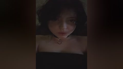 Video of a young woman with pale skin, dark hair, and a black strapless dress. She has a serious expression and wears a choker necklace. The background is dimly lit.