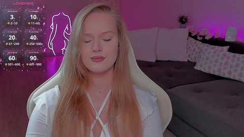 Video of a blonde woman with closed eyes, wearing a white top, sitting on a gaming chair, in a dimly lit room with a bed, pillows, and purple lighting.