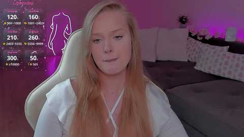 Video of a blonde woman with long hair, wearing a white top, sitting on a gaming chair in a dimly lit bedroom with a bed and purple lighting.