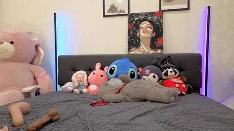 A video of a cozy, plush-filled bedroom with a gray headboard, stuffed animals, and a framed art piece with a serene woman's face.