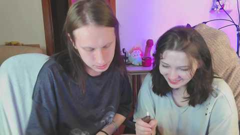 Video of two young women with pale skin, one with shoulder-length brown hair, the other with shoulder-length black hair, wearing casual clothes. They are seated on a light blue couch in a dimly lit room, holding a small, red, heart-shaped toy.