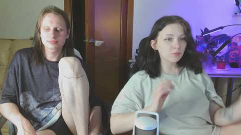 Video of a pale, slim, androgynous person with long, straight brown hair, wearing a dark shirt and light pants, seated beside a tattooed, dark-haired woman in a gray shirt. They sit in a dimly lit room with a wooden door and a blue-lit desk behind them.