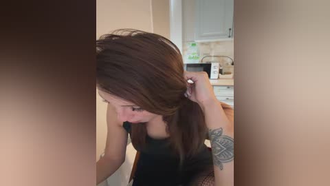Video of a young woman with long brown hair, wearing a black top, brushing her hair in a kitchen. She has a tattoo on her left wrist.