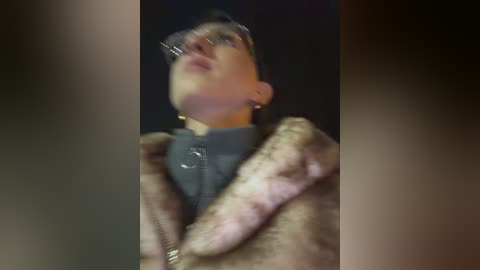 A blurry video of a person with glasses, a dark jacket, and a leopard-print coat, standing against a dimly lit background. The image is grainy and out of focus, making the subject indistinct.