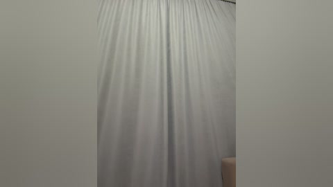 A video of a simple, empty room with a plain white curtain hanging vertically in the center. The curtain is slightly crumpled, creating a textured, wavy pattern. The background wall is a neutral off-white, and there is a beige cushion visible in the bottom right corner.