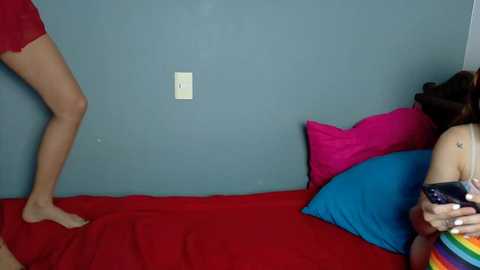 Video of a woman in a red dress standing on a red bed with a blue pillow, holding a phone, against a gray wall.