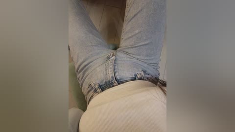 Video of a person sitting with legs crossed, wearing light blue jeans and a beige sweater, against a blurred background.