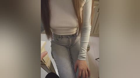 Video of a woman in a light grey, long-sleeved top and high-waisted, light blue jeans, sitting on a bed with a beige headboard, and a wooden dresser in the background.