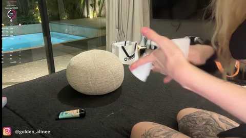 Video of a woman with long blonde hair painting a beige sphere on a black table, with a blue pool and greenery visible through a window.