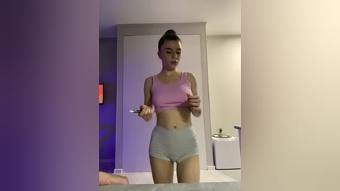 A video of a young Asian woman with a slim build, wearing a pink sports bra and grey shorts, adjusting her bra in a dimly lit bathroom.