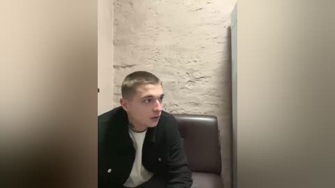 Video of a young, fair-skinned, short-haired man in a black jacket and white shirt, sitting in a dark chair against a light beige brick wall. He looks thoughtful, with a serious expression.