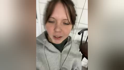 A video of a young Caucasian woman with fair skin and brown hair, wearing a gray hoodie, leaning against a glass door with headphones on, looking slightly surprised.