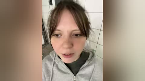 A candid video of a young Caucasian girl with light skin, brown hair, and light-colored hoodie, taken from a low angle, indoors with white tiled walls in the background.