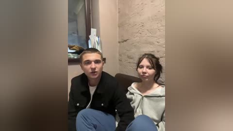 A video of a young woman and man sitting side-by-side in a dimly lit, beige-walled room. The woman, with dark hair, wears a hoodie, and the man, with short hair, sports a black jacket and jeans.