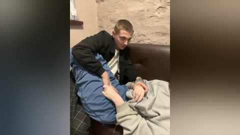 A video depicts a young man in a black jacket, blue overalls, and a striped tie, helping a seated elderly man in a grey shirt and beige blanket, in a rustic room with a brown leather couch and stone wall.