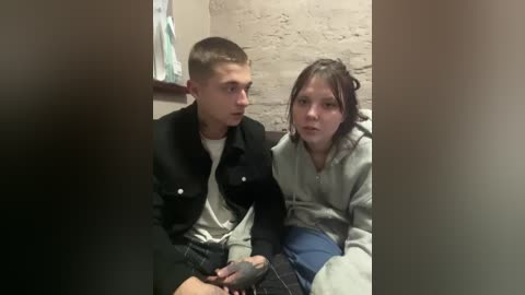 A video of a young Caucasian boy with short hair in a black jacket and a girl with shoulder-length brown hair in a gray hoodie, sitting side-by-side in a bathroom with a brick wall and hanging towels.