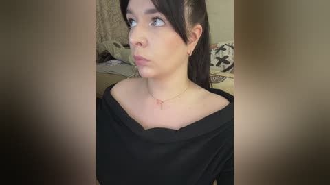 Video of a young woman with fair skin, dark hair, wearing a black off-the-shoulder top, standing in a bedroom with a bed and patterned pillows in the background.